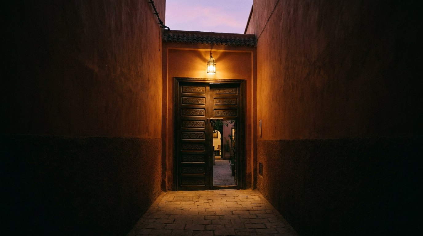 Façade du riad au crépuscule, porte cintrée entrouverte, lanternes de cuivre allumées, ruelle de la Médina. Format vertical 3:4.
